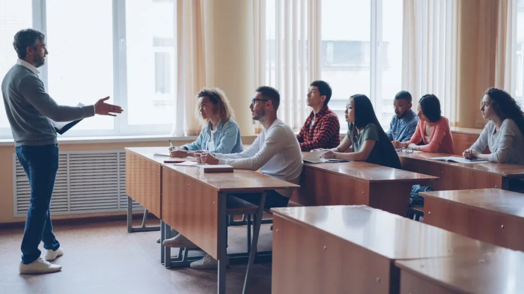 A group of adult students sitting at wooden desks in a bright classroom, listening to a male instructor at the front.