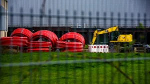 Construction equipment and large red utility pipes at a fenced-off roadworks site.