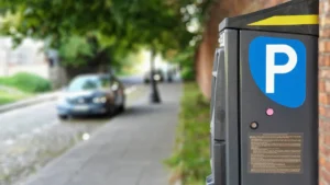 A close-up of a street parking payment machine with a blue and white parking sign, with a car blurred in the background.