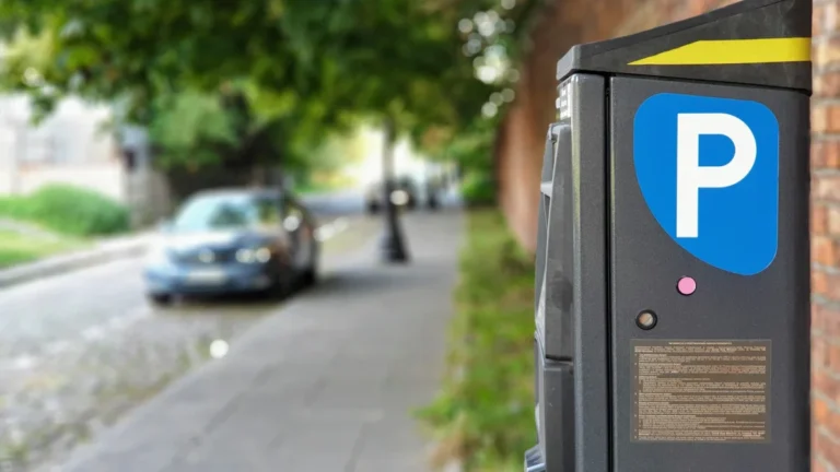 A close-up of a street parking payment machine with a blue and white parking sign, with a car blurred in the background.