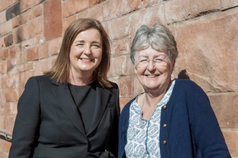 Two smiling women stand side-by-side in front of a textured red stone brick wall.