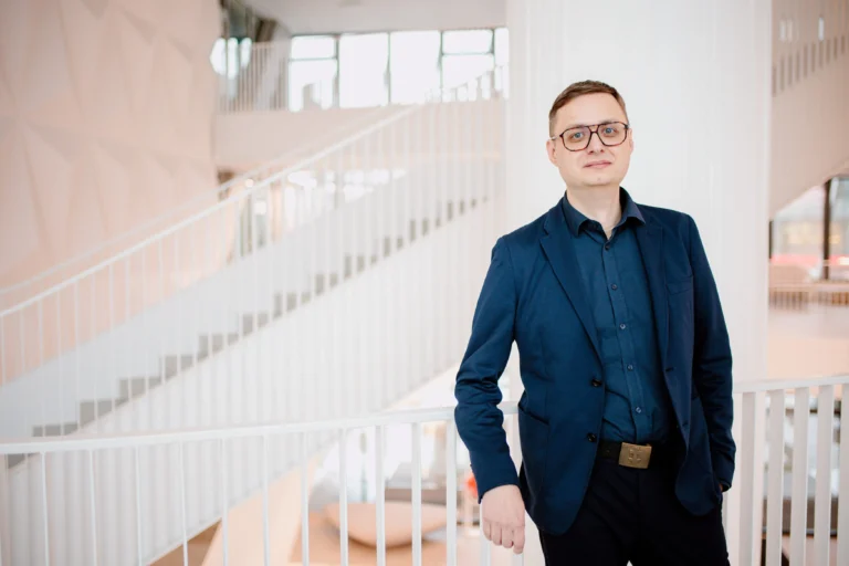Man wearing a navy blazer and glasses standing in a bright, modern interior with white stairs.
