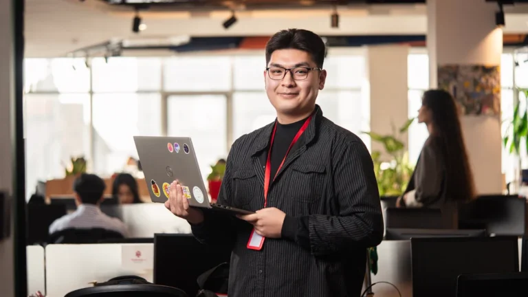 A tech professional holding a laptop and smiling in a busy modern office setting.
