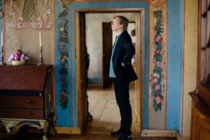 A man in a black blazer stands in a doorway surrounded by ornate floral wall murals.