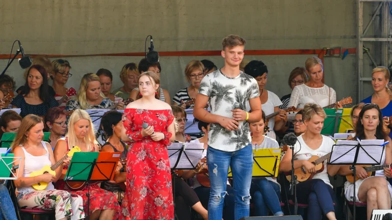 A young man and woman singing on a festival stage accompanied by a large orchestra.