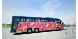 A grey bus featuring a pink wrap with numerous pug illustrations parked in an open lot.