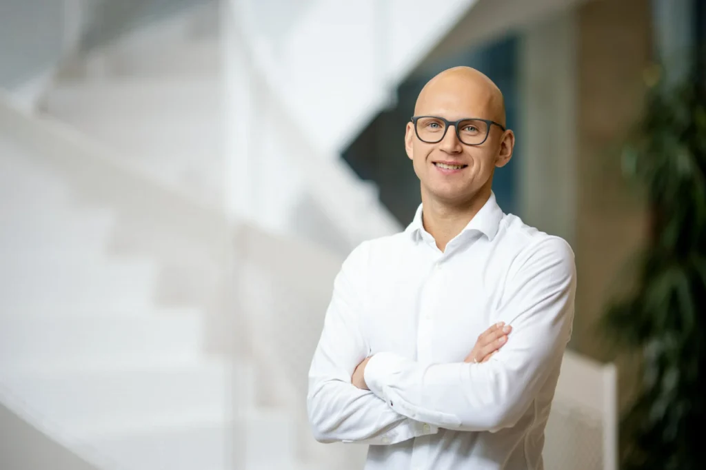 Smiling bald man with glasses and arms crossed wearing a white shirt against a white staircase.