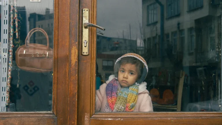 A young child in a winter scarf looks thoughtfully through a window reflecting urban buildings.