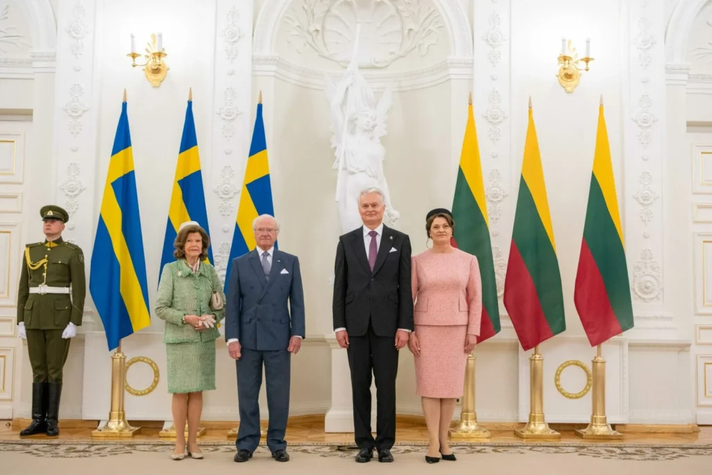 Four people in formal attire stand between Swedish and Lithuanian flags in an ornate hall.
