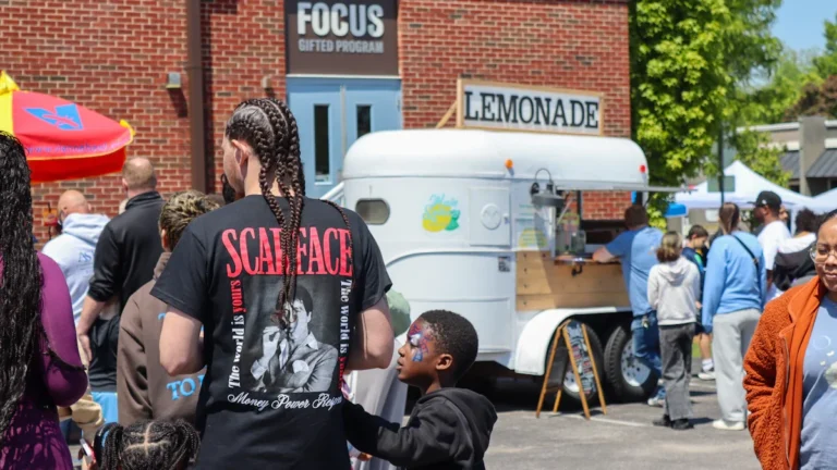 A diverse group of community members gathered at a sunny outdoor event featuring a 'Lemonade' stand and the 'FOCUS Gifted Program' building.