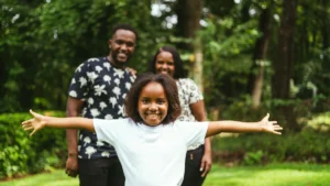 A joyful young girl with her arms wide open, standing in front of her smiling parents in a lush green park.