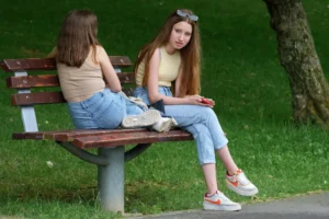 Two teenage girls wearing tank tops and jeans sit on a wooden park bench outdoors.