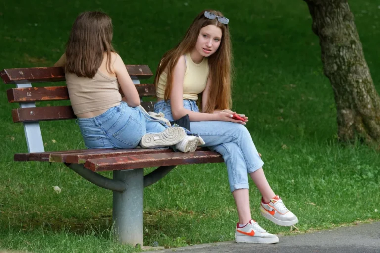Two teenage girls wearing tank tops and jeans sit on a wooden park bench outdoors.