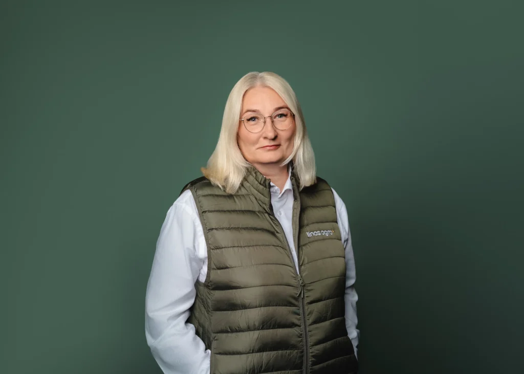 A blonde woman in glasses wearing an olive green quilted vest against a dark green background.