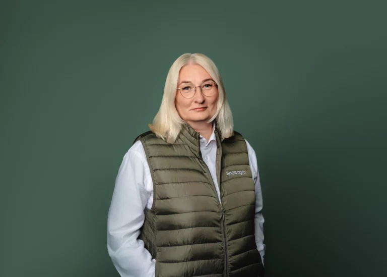 A blonde woman in glasses wearing an olive green quilted vest against a dark green background.