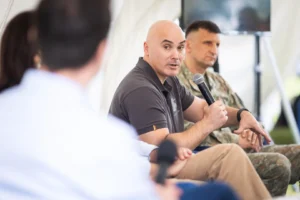 A bald man in a dark polo shirt speaks into a microphone during a panel discussion.