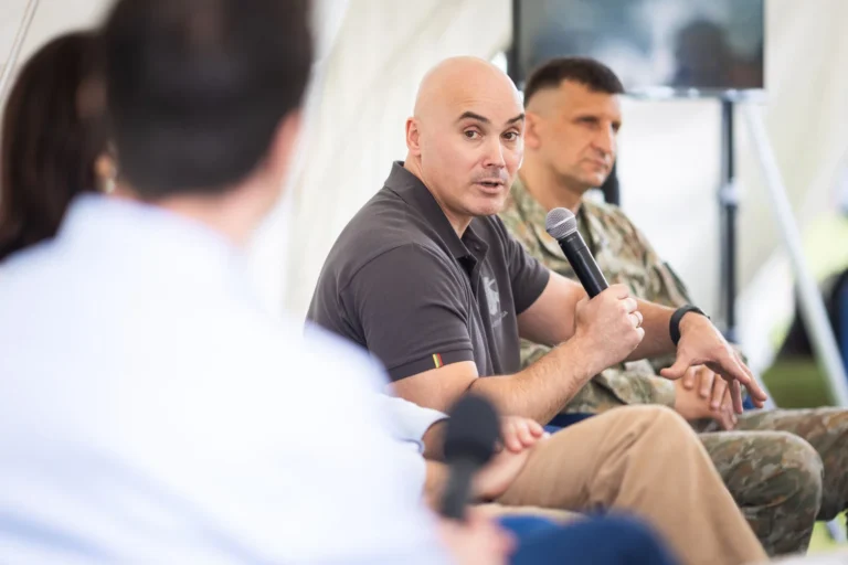 A bald man in a dark polo shirt speaks into a microphone during a panel discussion.