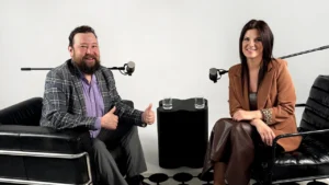 A bearded man and a woman in blazers smile while sitting in a minimalist podcast studio.