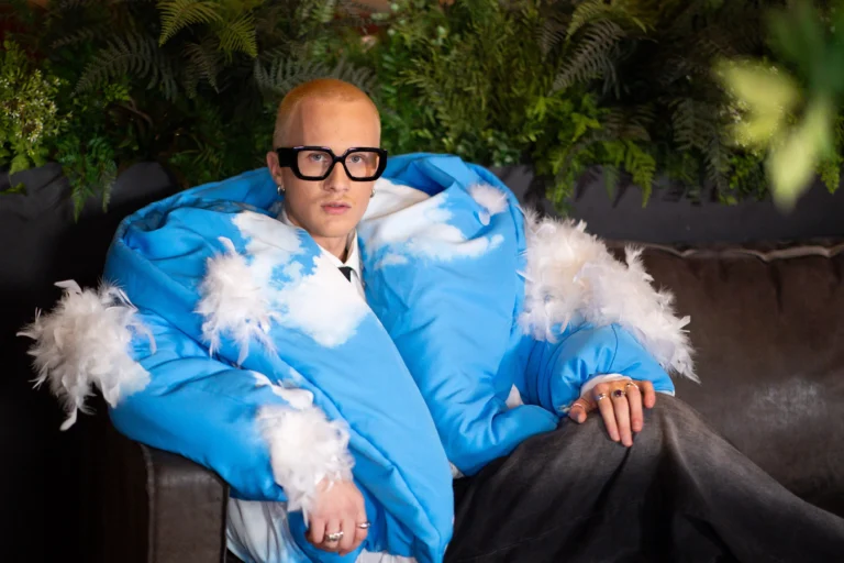 Blonde man in an oversized blue cloud-pattern jacket with white feathers sitting on a couch.