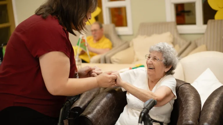 A caregiver in a maroon shirt assists an elderly woman using a walker in a care setting.