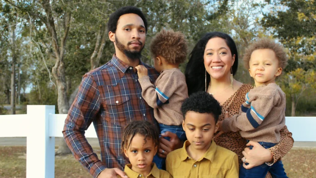 A diverse family of five with three young children posing together outdoors near a white fence.