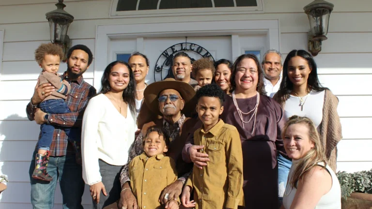 A large, diverse multi-generational family stands together smiling in front of their home, representing Brent foster carers and their community impact.