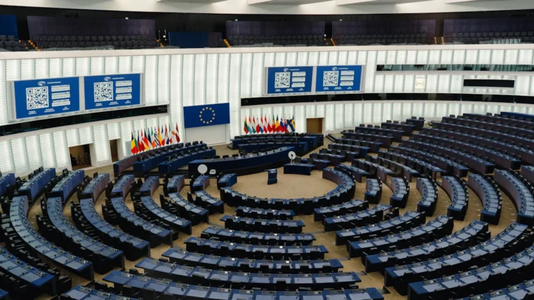 An empty legislative assembly chamber with rows of blue desks arranged in a semicircle.