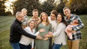 A large, joyful family stands together in a field, with a young child and baby present.