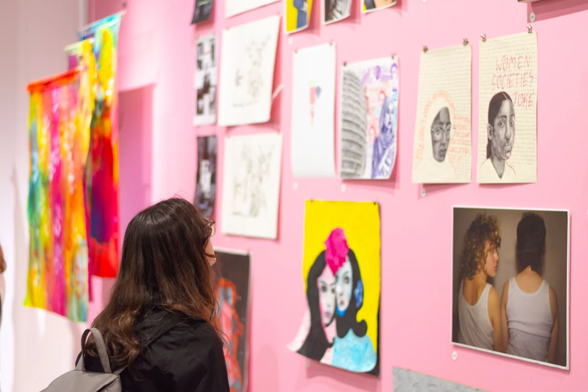 A woman with long dark hair views various art prints on a pink wall.