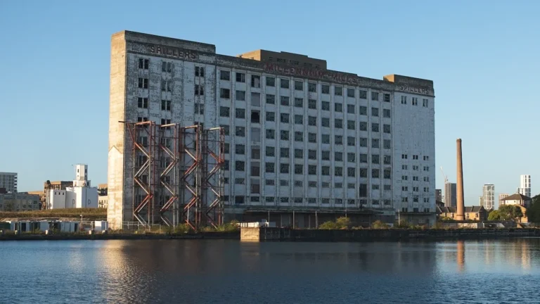 A large, historic white industrial warehouse building reflected in a body of water under a clear blue sky.