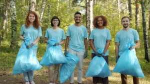 A diverse group of five Cheshire East volunteers in matching blue t-shirts standing together in a forest, holding full blue litter bags and smiling.