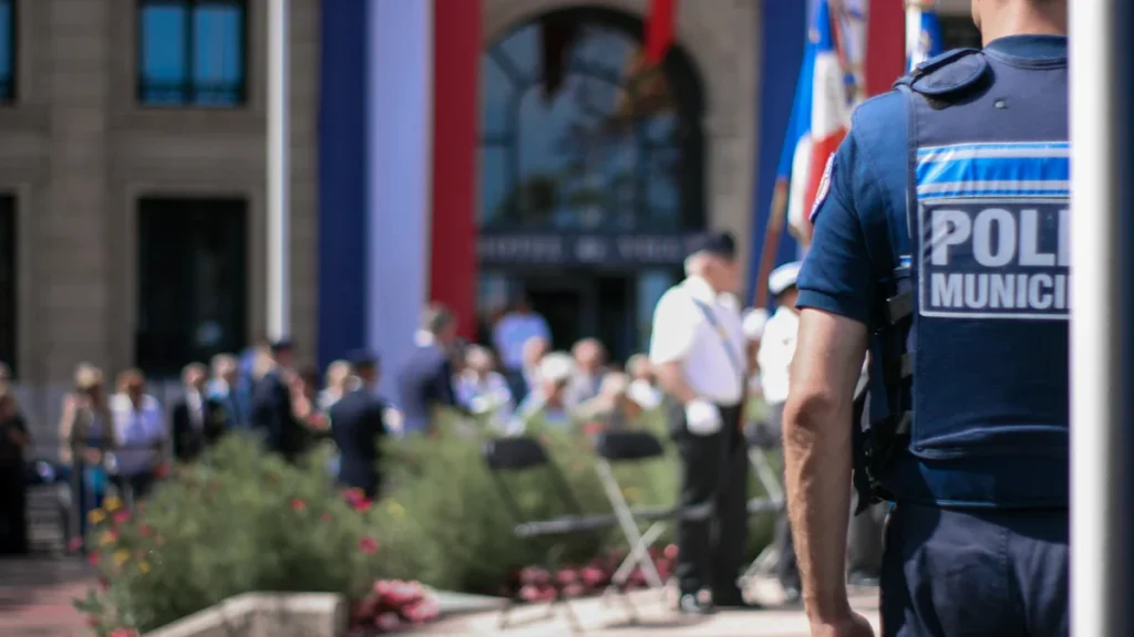 A blurred municipal building with flags and a police officer overseeing a public civic ceremony.