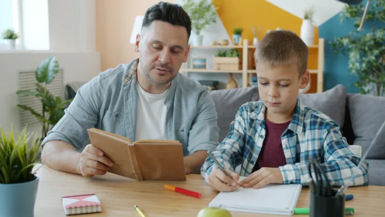 A father helps his young son with reading and writing at a desk, representing literacy support at home.