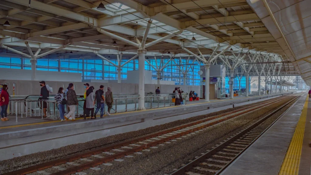 A modern, wide-span train station platform with a high white steel roof structure and residents waiting for transit.
