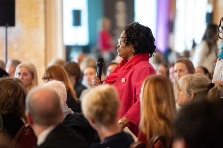 Black woman in a pink jacket holding a microphone while standing among a seated audience.
