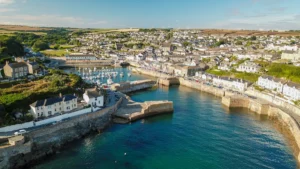 An aerial view of a coastal town in Cornwall featuring a harbor and densely packed residential houses under a clear blue sky.