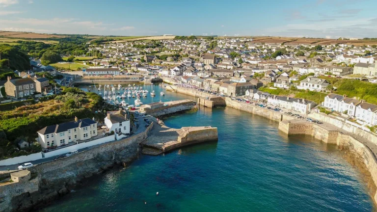 An aerial view of a coastal town in Cornwall featuring a harbor and densely packed residential houses under a clear blue sky.