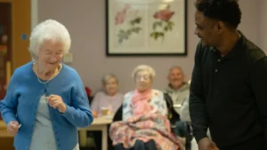 An elderly woman in a blue cardigan dances with a caregiver in a UK community center.