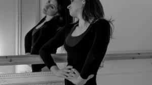 A woman in black athletic clothing stands at a barre in a mirror-lined fitness studio.