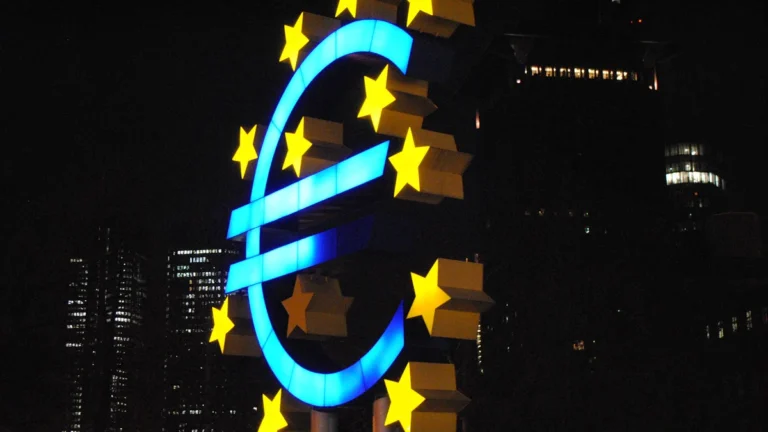 A large illuminated blue Euro symbol sculpture with yellow stars at night against a city backdrop.