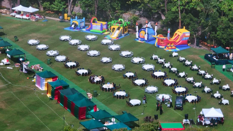 Aerial view of a large-scale outdoor community event at a park featuring numerous banquet tables, gazebos, and inflatable bouncy castles.