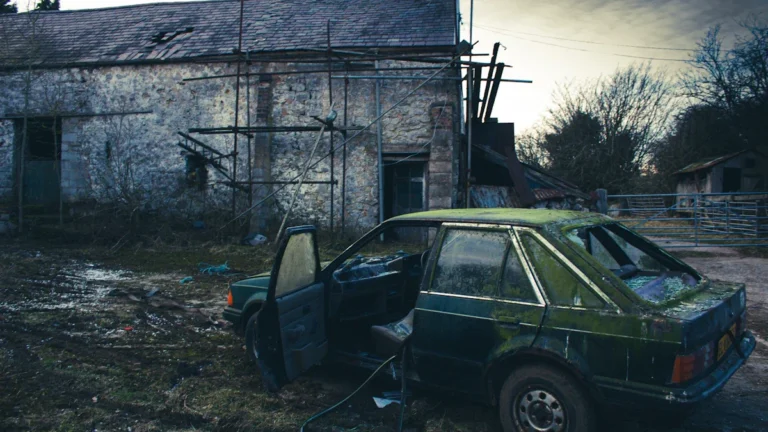 A green hatchback covered in moss and grime sits abandoned near a stone building in the UK.