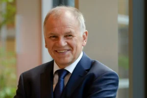 Portrait of a smiling older man with grey hair wearing a navy suit and tie.