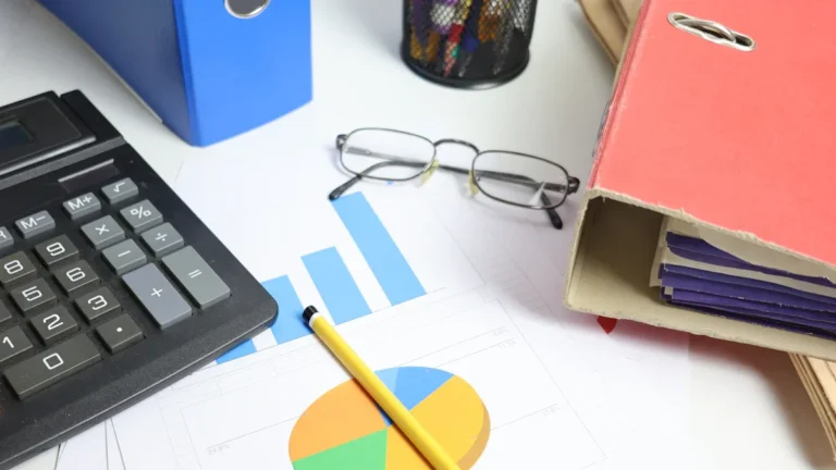 A business desk featuring a calculator, financial charts, eyeglasses, and office binders for data reporting.