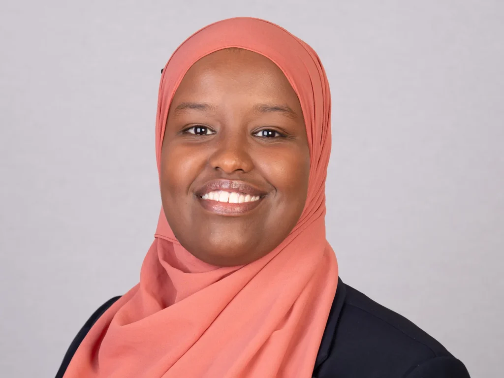 Close-up portrait of a smiling Black woman wearing a coral hijab and dark blazer.