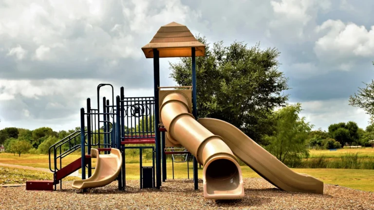 A modern tan and brown children's playground structure with slides in a green public park setting.