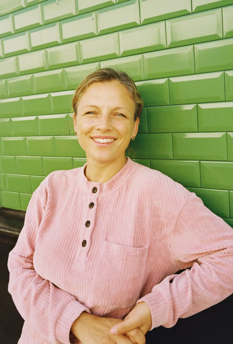 A smiling woman in a pink corduroy shirt stands against a green tiled wall.