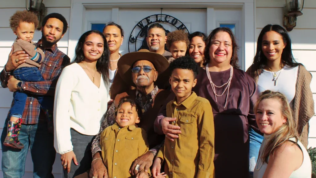 A large, multi-generational family of twelve people posing for a portrait in front of a white house, representing the 26-year legacy of fostering in Ealing.