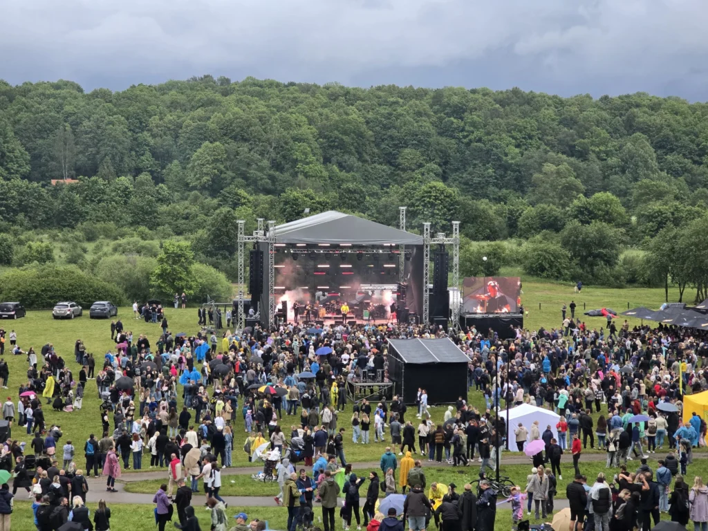 Crowd watches an outdoor concert stage in a grassy field backed by a dense green forest.