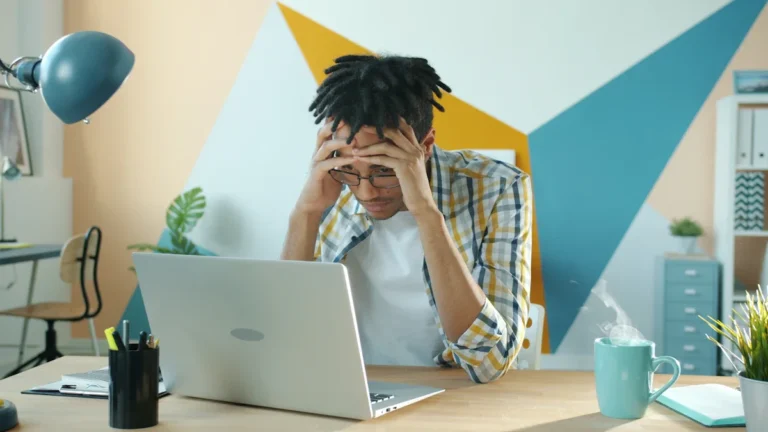 A stressed young Gen Z man sitting at a laptop in a modern workspace, illustrating the struggle with financial crisis management despite having technical skills.